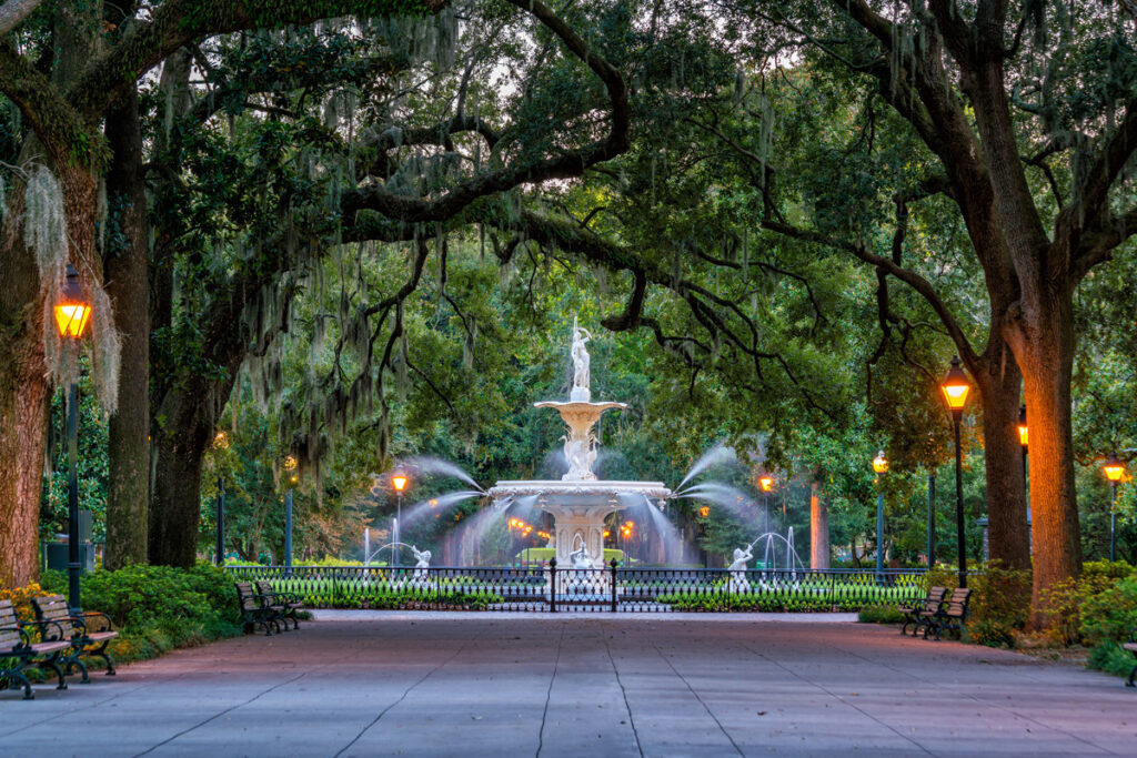 Twilight view of a tranquil park with a central fountain surrounded by illuminated street lamps and ancient oaks.