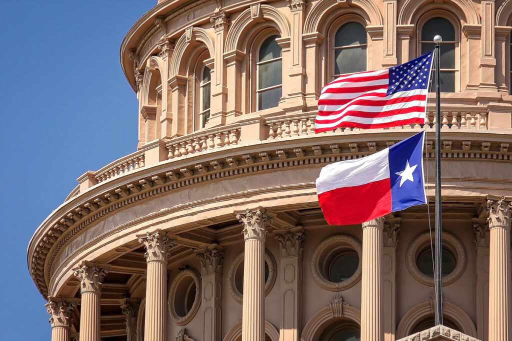 American and Texas state flags waving in front of an ornate building, symbolizing state and national pride.