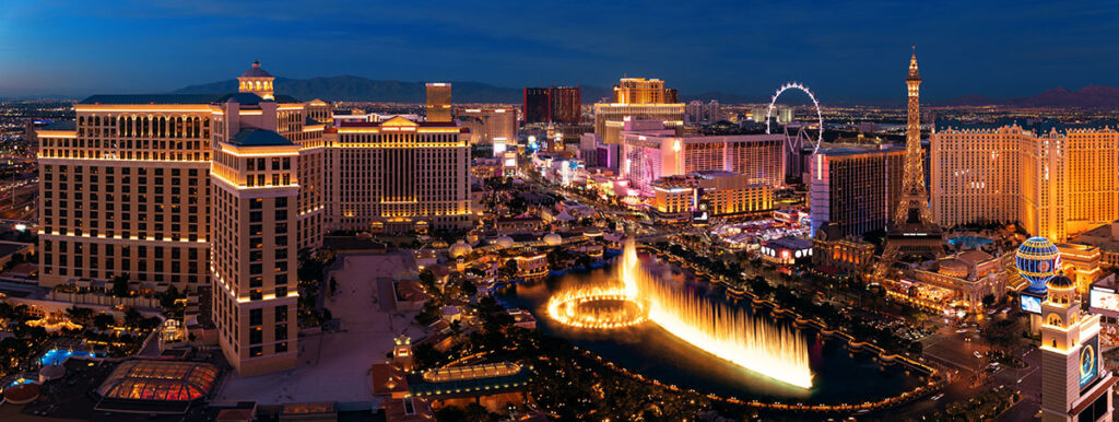 Panoramic view of the Las Vegas Strip at twilight, showcasing the Bellagio Hotel's fountain show, with a vibrant display of lights and casinos against a mountainous backdrop.